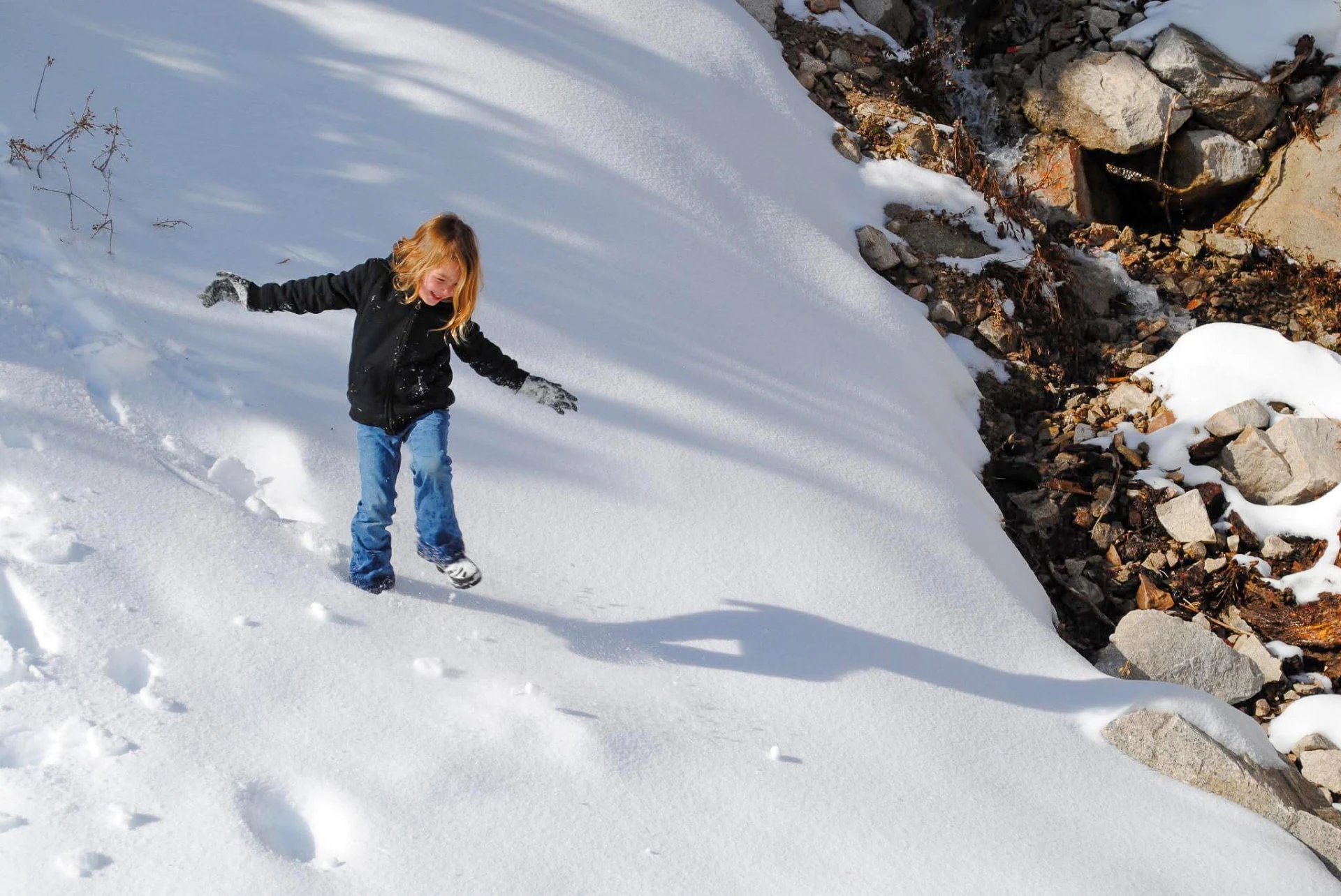 A young girl in a dark jacket and jeans playing on a snowy hillside