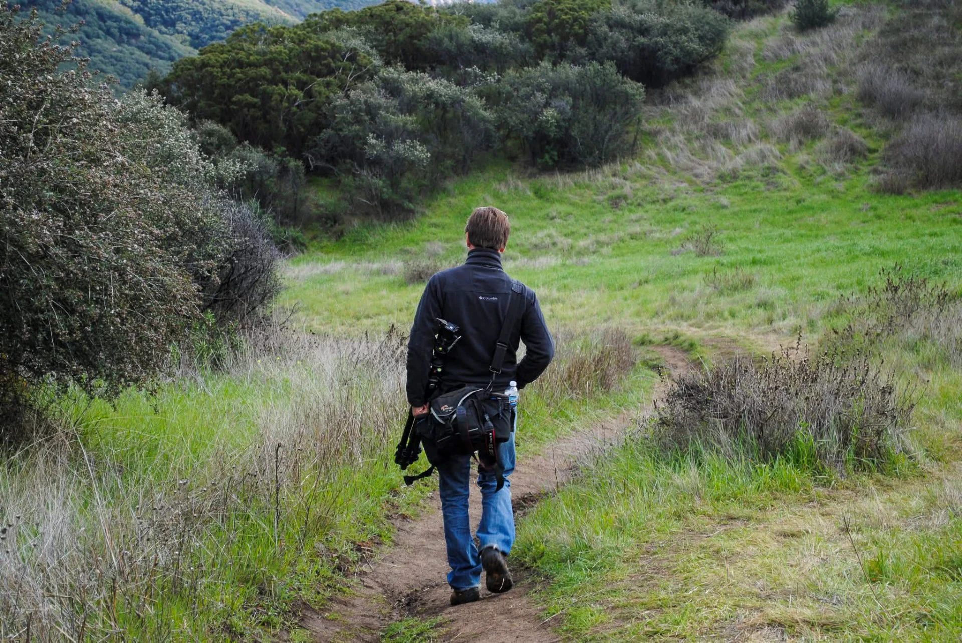 A photographer walking along a narrow green trail between wooded hills